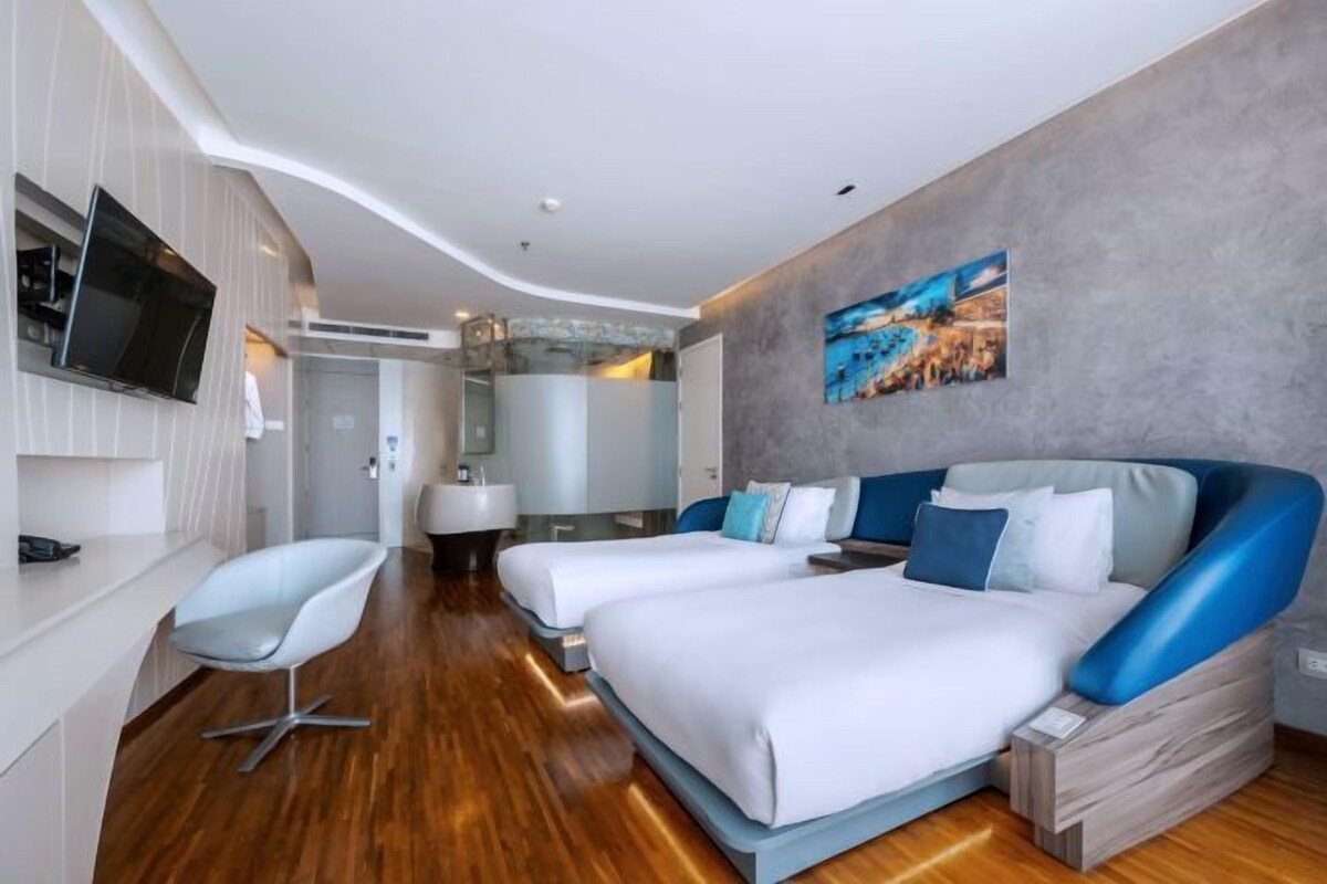 A modern hotel room with two beds, a TV mounted on the wall, a bathroom with a glass shower, and a painting on the wall.