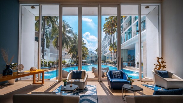 A modern hotel room with a large glass door leading to a pool area, featuring a view of palm trees and a multi-story building. - Hotel Baraquda Pattaya By Heeton  (Pattaya)