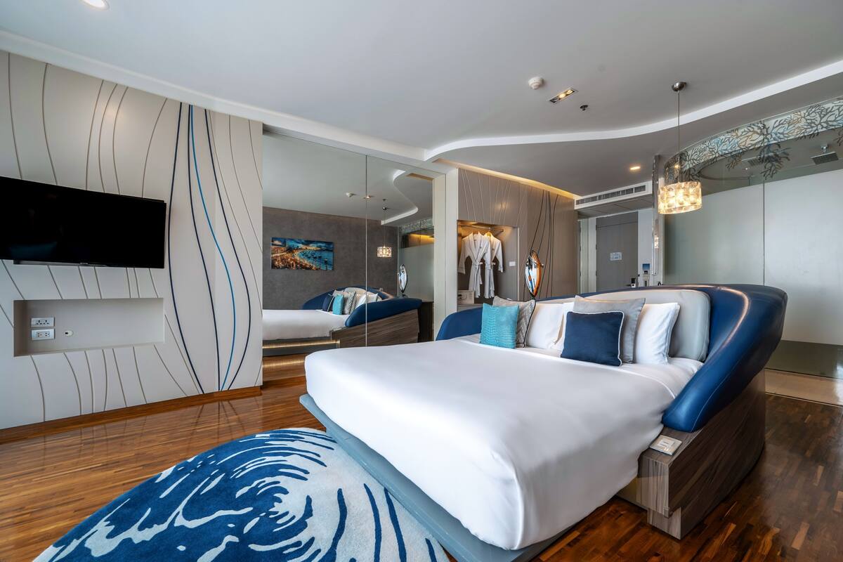 Una habitación de hotel moderna con techo curvo, una cama grande con ropa de cama azul y blanca, un televisor de pantalla plana montado en la pared y un cuadro en la pared.