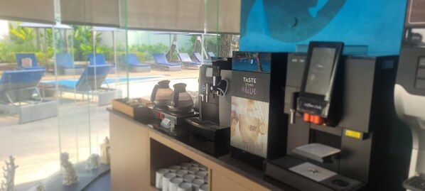 A coffee machine with a screen displaying "TASTE COMES alive" and a view of a poolside area with lounge chairs.