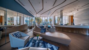 A modern lounge area with sofas and armchairs, a reception desk, and large windows offering views of palm trees and a pool. - Hotel Baraquda Pattaya By Heeton  (Pattaya)