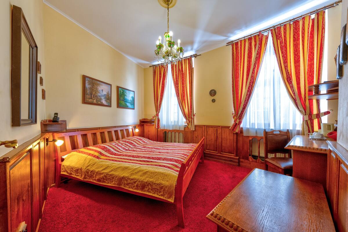 A traditional room with a wooden bed, a desk, and a chair. There is a chandelier, a painting on the wall, and curtains on the windows.