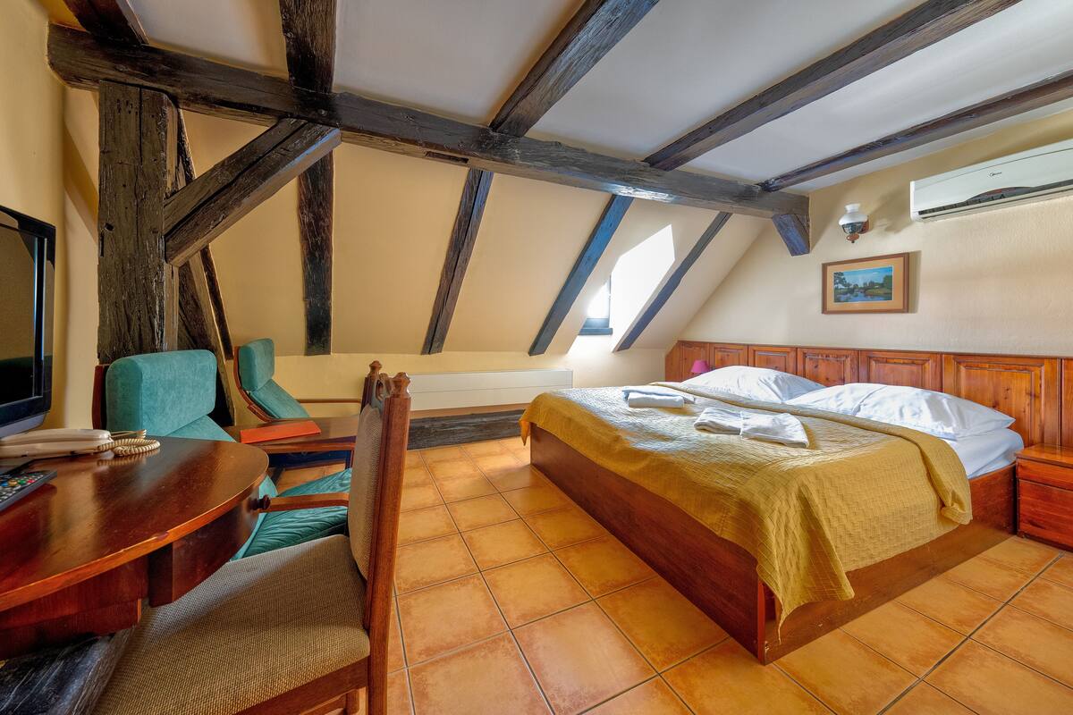 A room with a wooden ceiling, a bed with a yellow bedspread, a wooden desk with a chair, and a television on the left.