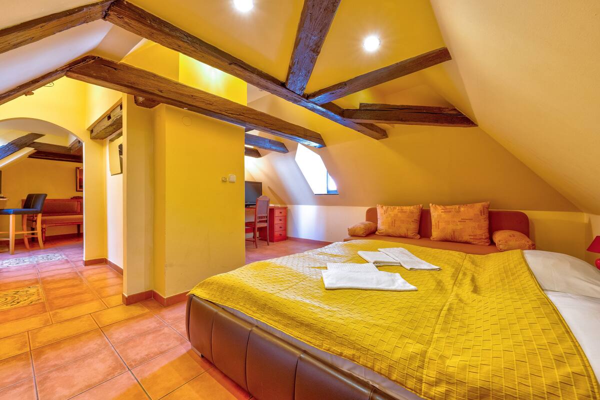 A bedroom with a high ceiling, exposed wooden beams, a yellow bedspread, and a view of the adjacent dining area.