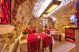 Un restaurante con paredes de piedra, manteles rojos y sillas de madera. Las mesas están dispuestas en forma de U y hay un mostrador de madera con una caja registradora.