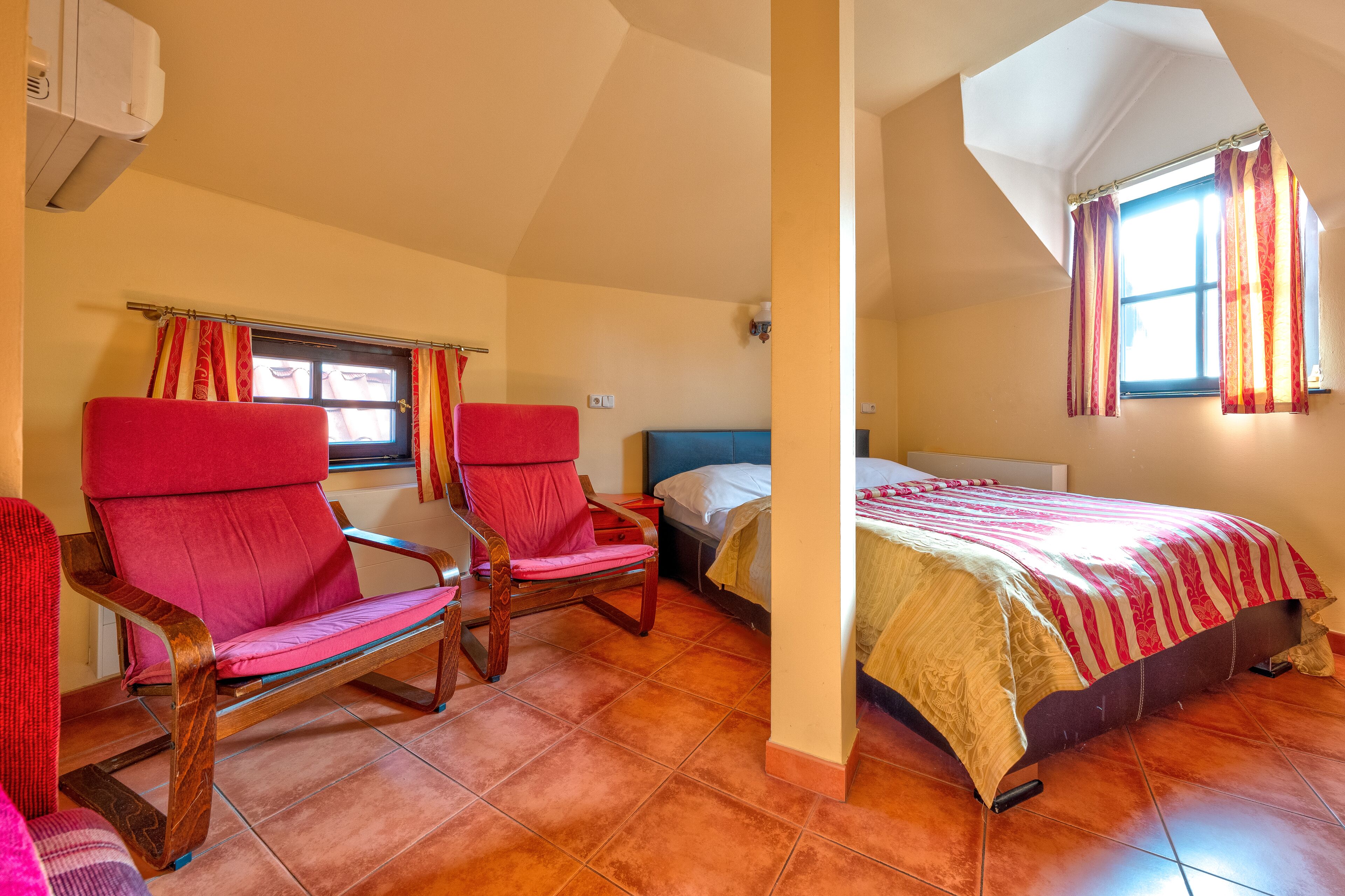 A bedroom with two red chairs, a bed with a striped bedspread, and a window with curtains.