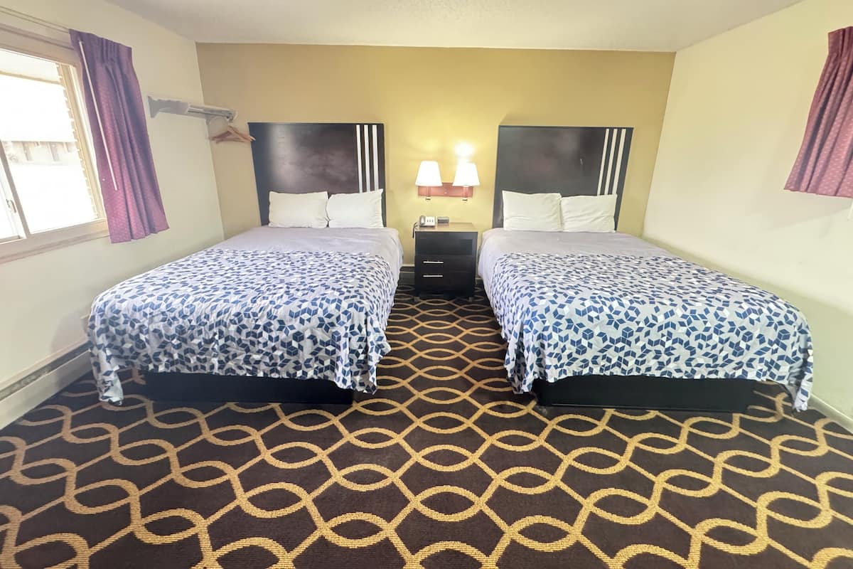 A hotel room with two beds, a patterned carpet, and a nightstand with lamps.