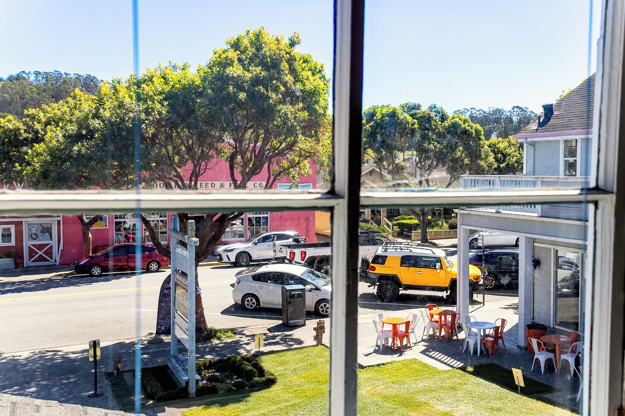 A view through a window of a street with parked cars, a tree, and a building with outdoor seating.