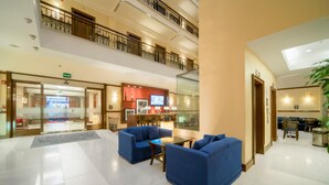 A hotel lobby with a glass partition, blue sofas, a reception desk, and a staircase to the upper level.