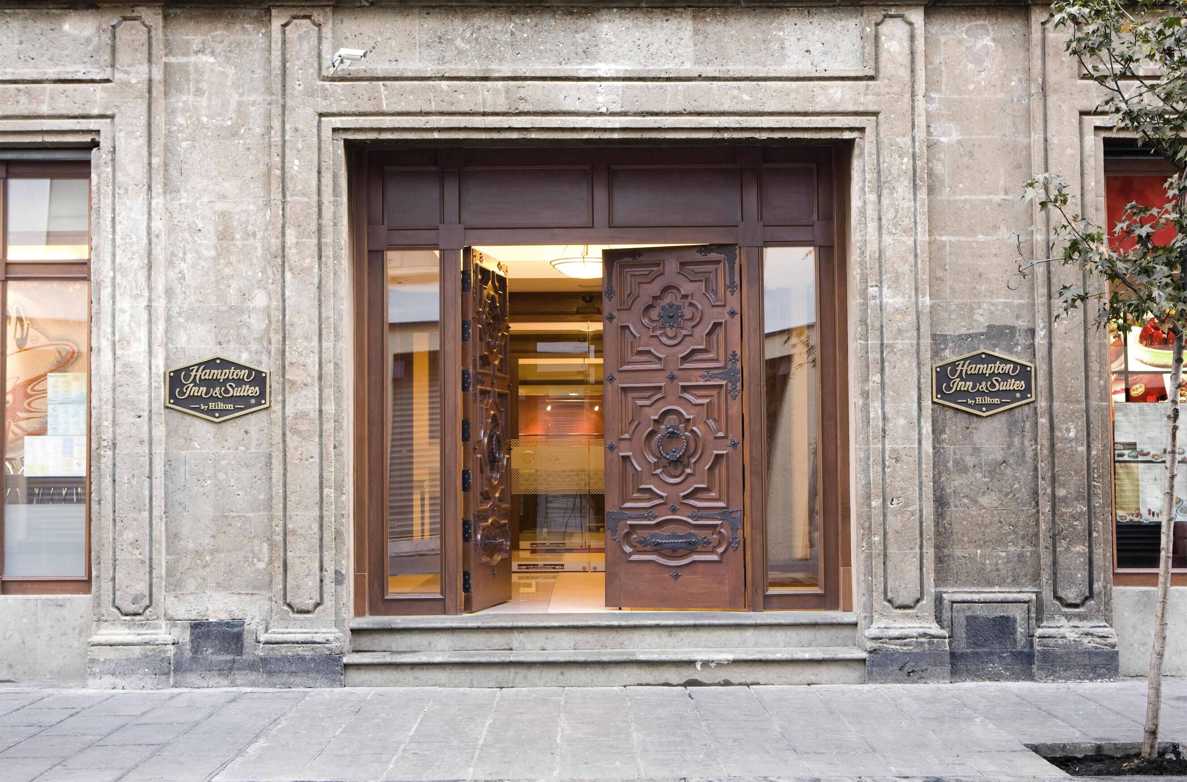A hotel entrance with a large wooden door, two signs indicating 'Hampton Inn & Suites by Hilton,' and a visible interior through the glass.