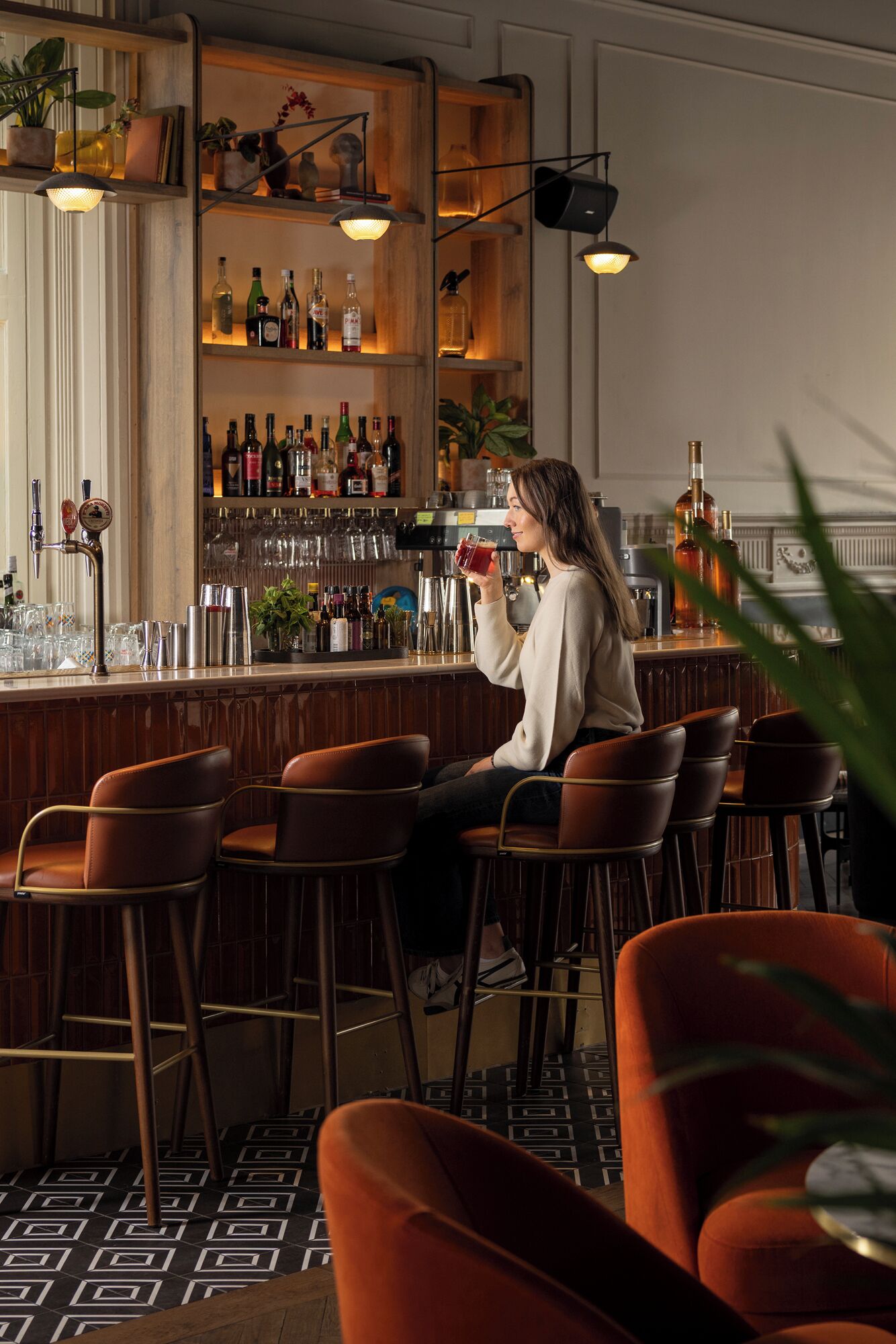 A bar area with a person holding a drink, shelves with bottles, and bar stools.