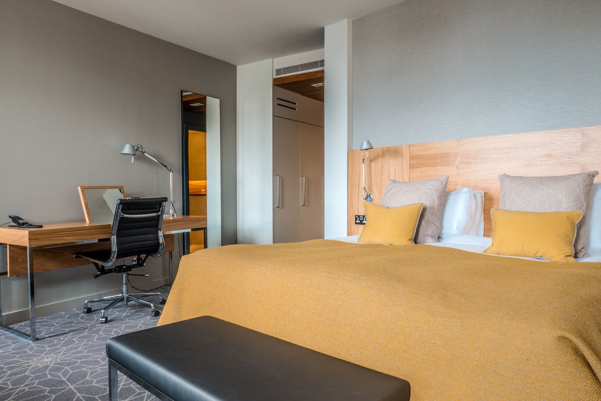 A hotel room with a bed, a desk, a chair, a bench, and a mirror.