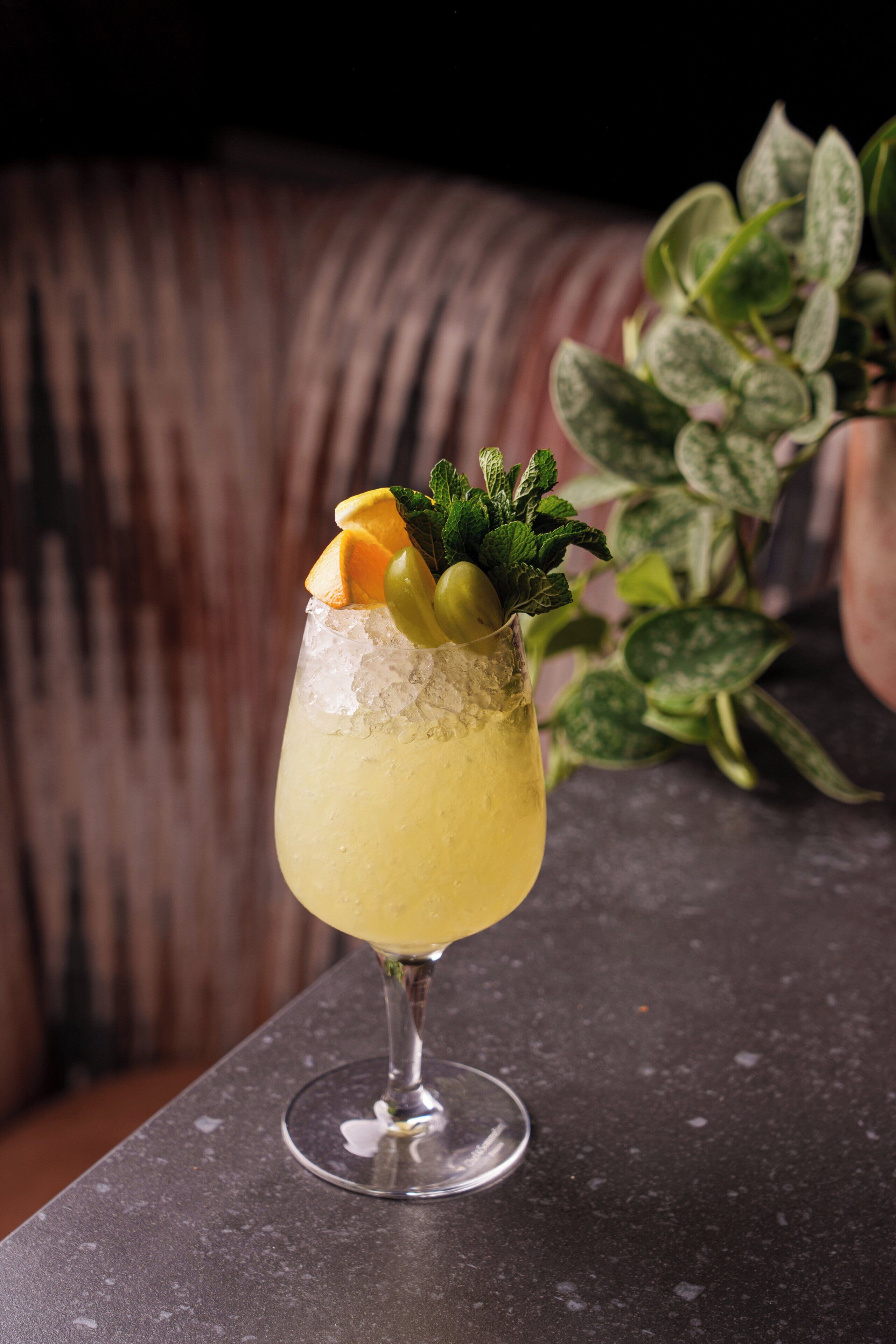 A cocktail glass with ice, garnished with mint, orange slice, and olives, placed on a dark surface.
