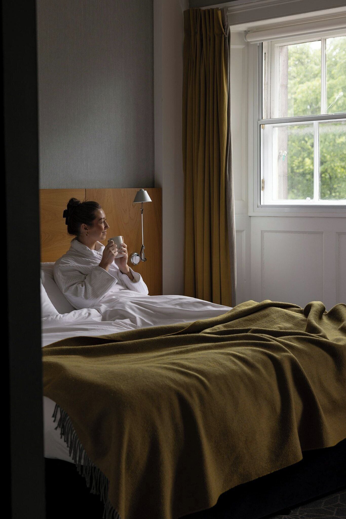 Een kamer met een bed, een bedlampje, een raam met gordijnen en uitzicht op groen.