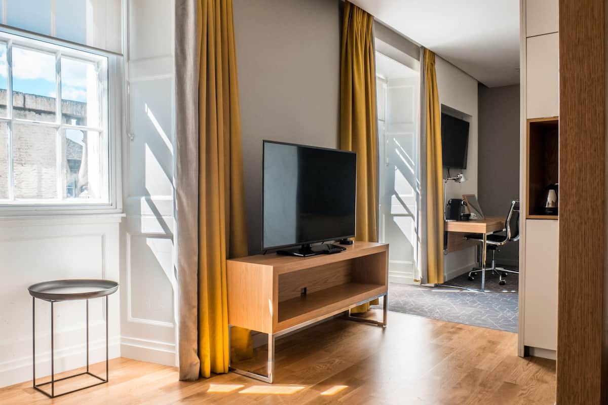 A modern living room with a flat-screen TV on a wooden stand, large windows with curtains, and a small side table.