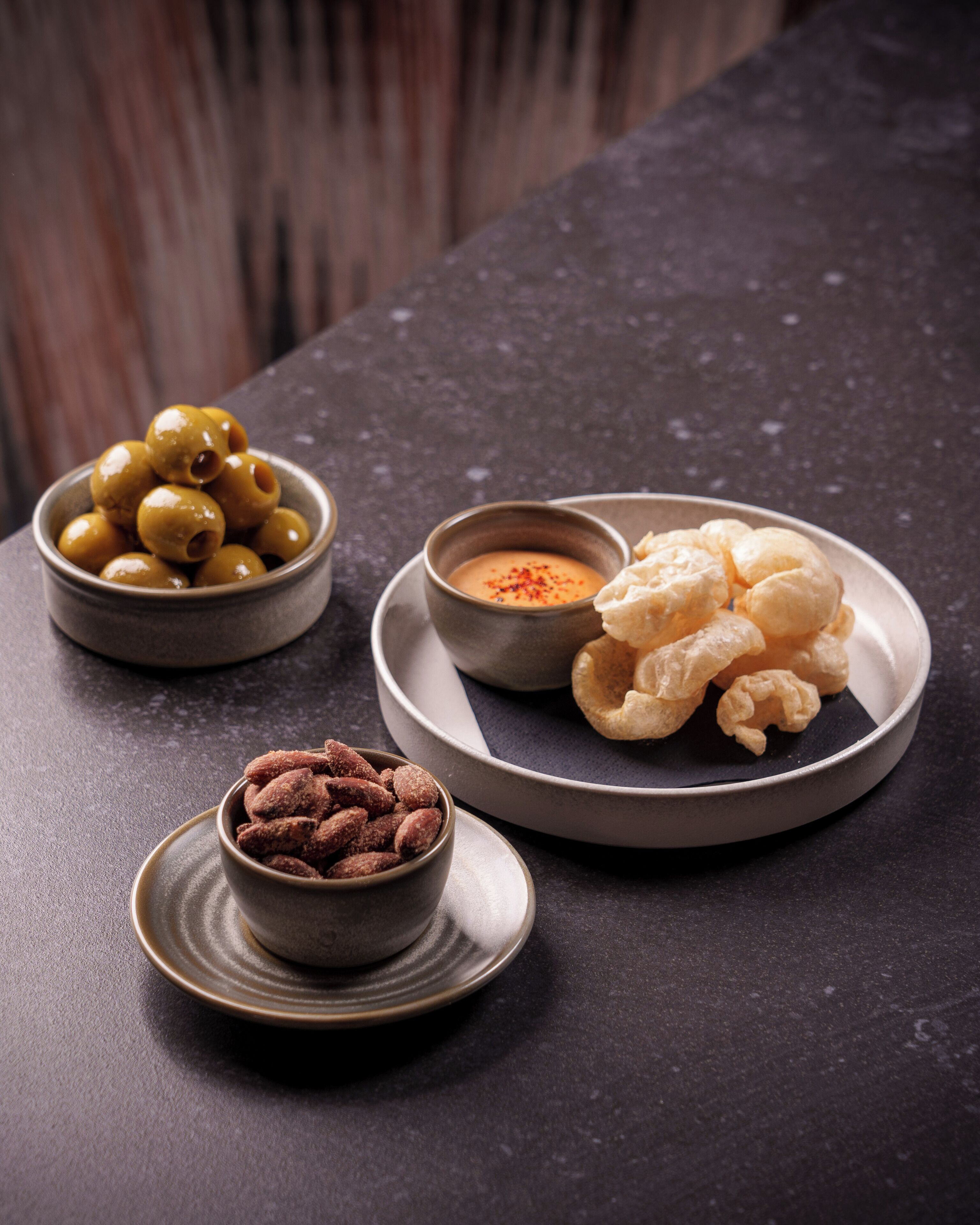 A table with three bowls containing olives, a creamy sauce, and a plate with pastries.