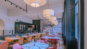 A spacious dining area with round tables and orange chairs, large hanging lamps, and a view of a building with a mural. - Apex Waterloo Place Hotel (Edinburgh)