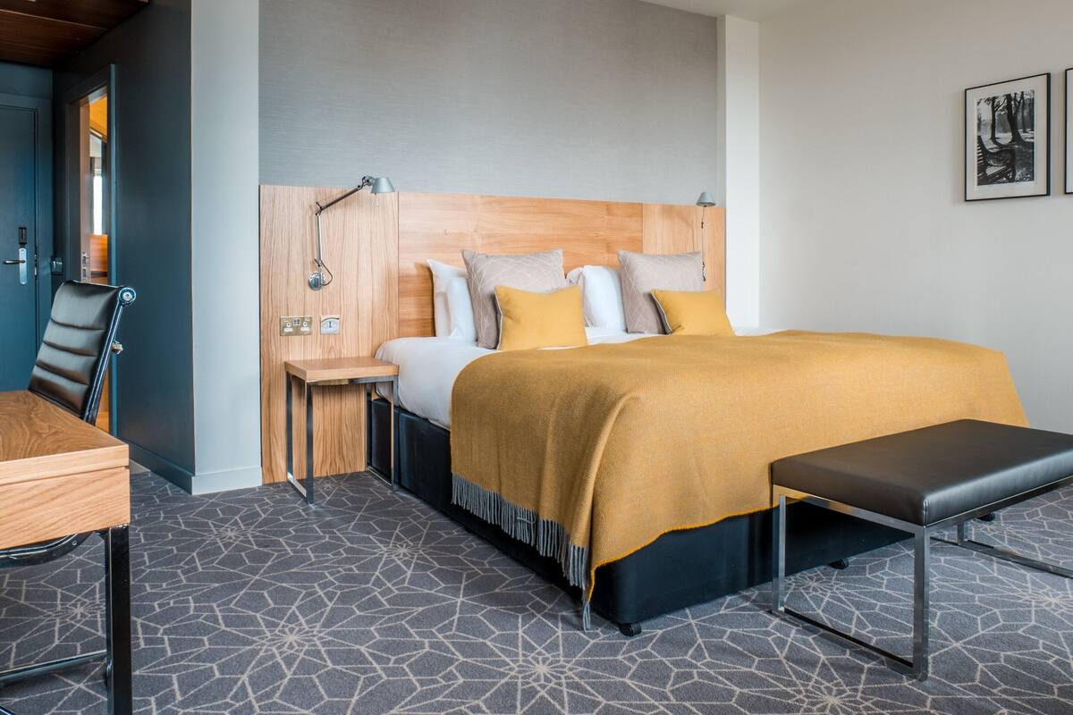 A modern hotel room with a wooden headboard, a bed with a mustard-colored blanket, a bedside table, a chair, and a bench.