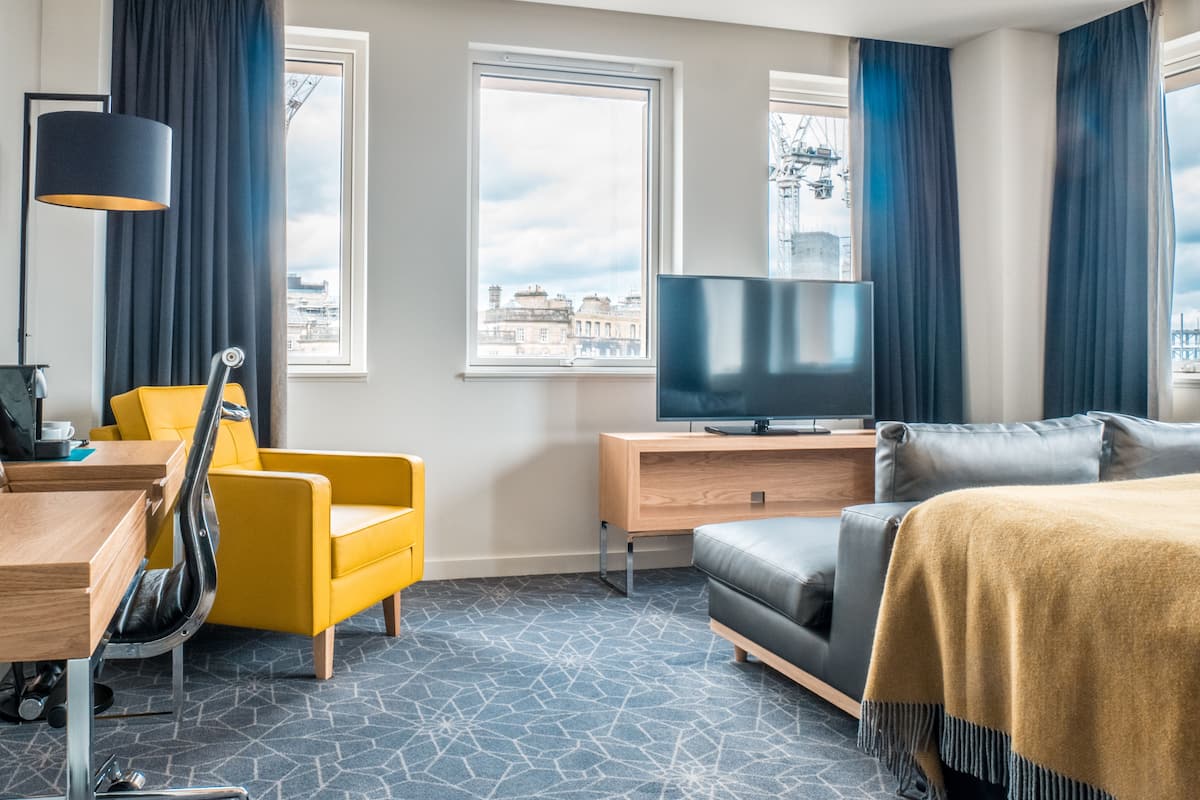A hotel room with a modern design, featuring a yellow armchair, a grey sofa, a flat-screen TV, and large windows offering a city view.