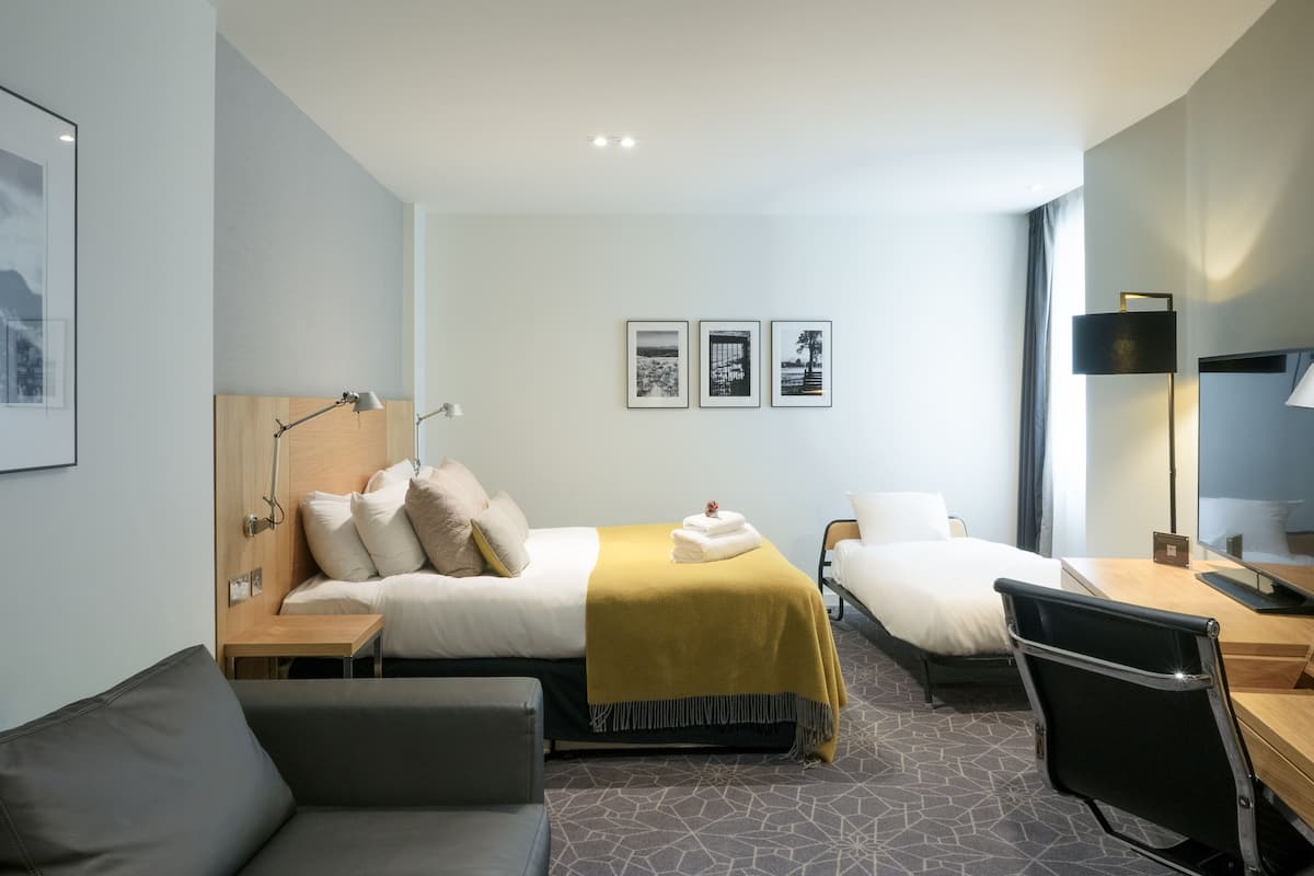 A modern hotel room with two single beds, a desk, a chair, a sofa, and a flat-screen TV.