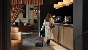 A woman in a coat pulling a suitcase, standing at a reception desk with a dark interior and modern pendant lights. - Apex Waterloo Place Hotel (Edinburgh)