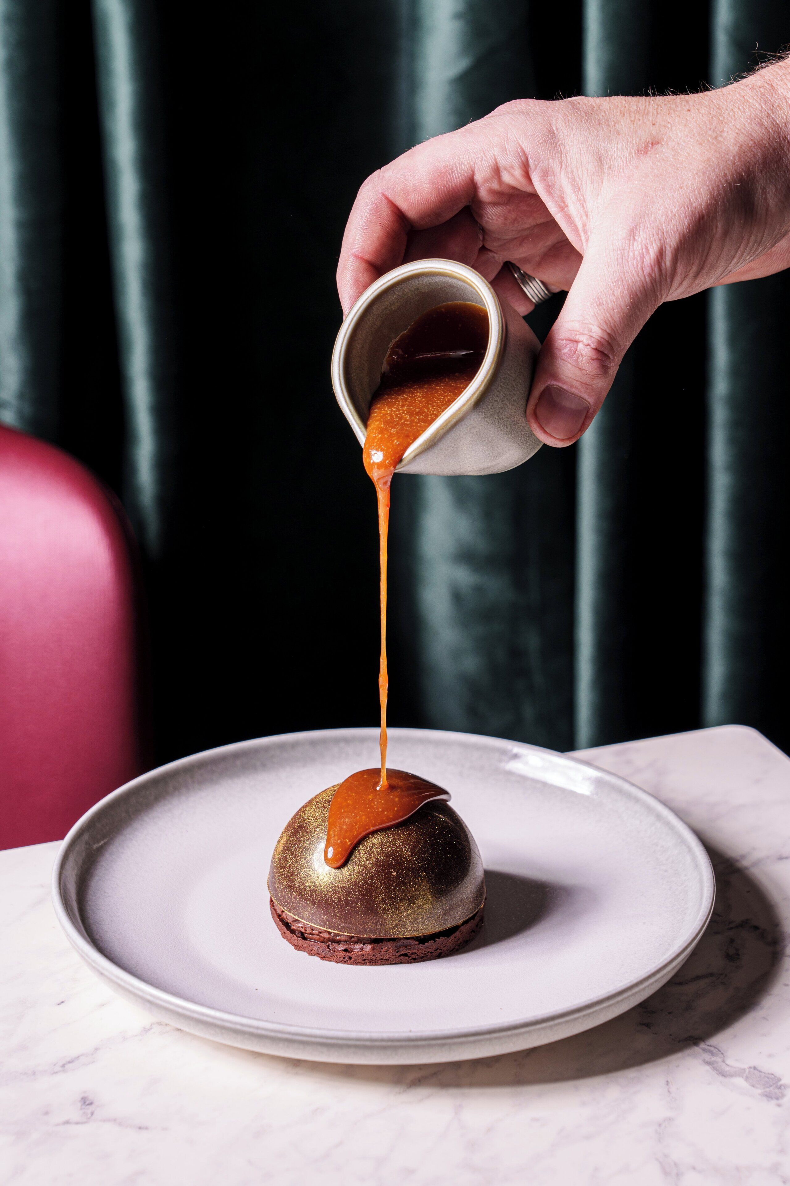 A hand pouring sauce over a dessert on a plate.