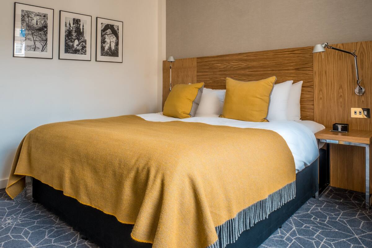 A neatly made bed with a mustard-colored blanket, a wooden headboard, and a nightstand with a lamp. There are three framed pictures on the wall.