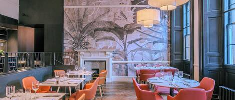 Interior de restaurante con piso de madera, sillas naranjas y rojas, mesas dispuestas con vasos y cubiertos, y un mural con follaje tropical.