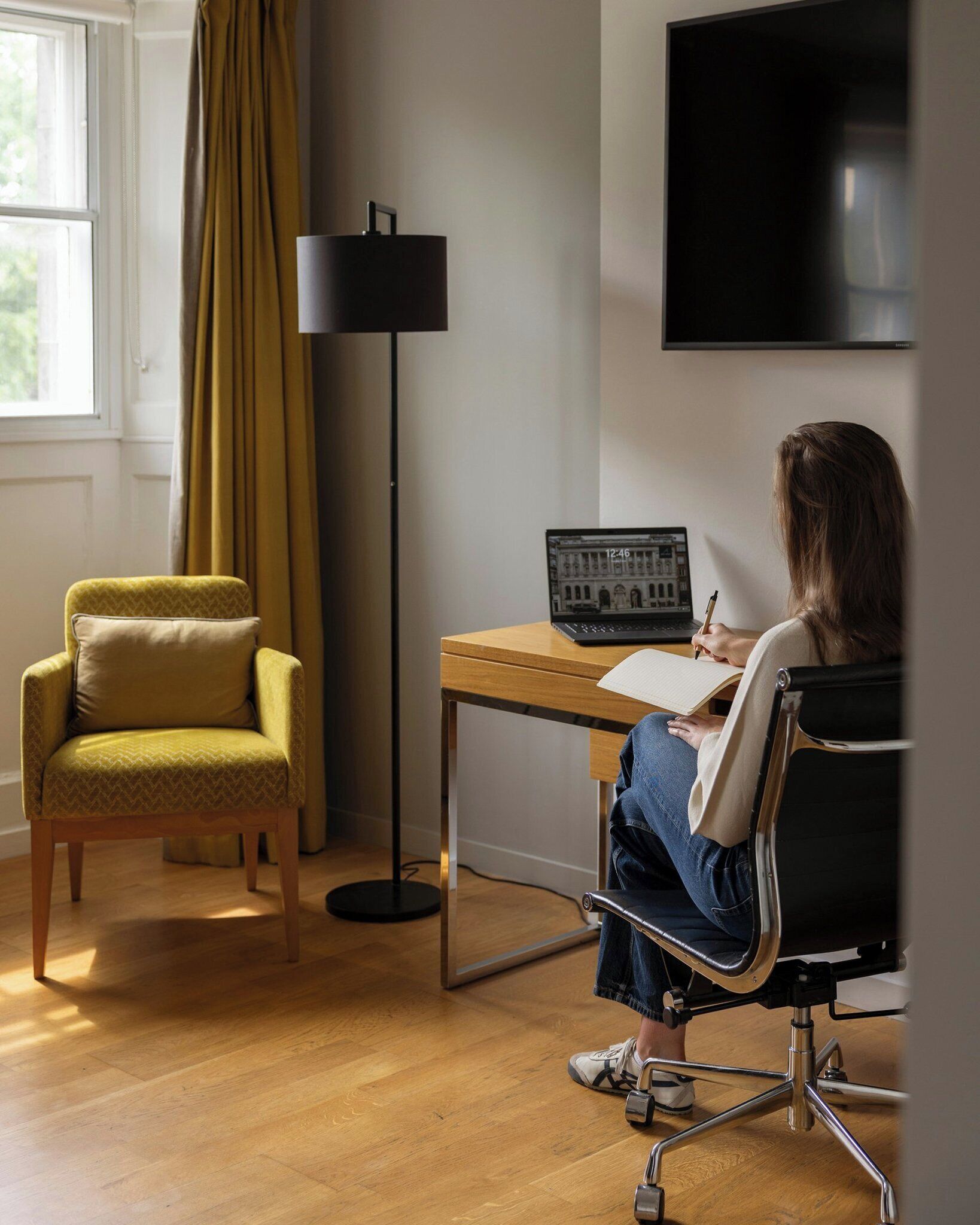 A room with a wooden desk, a laptop, a chair, a floor lamp, and a TV on the wall.