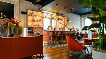 Una zona de bar con suelo de madera, taburetes rojos y una barra. Se ven estantes con botellas y plantas al fondo.