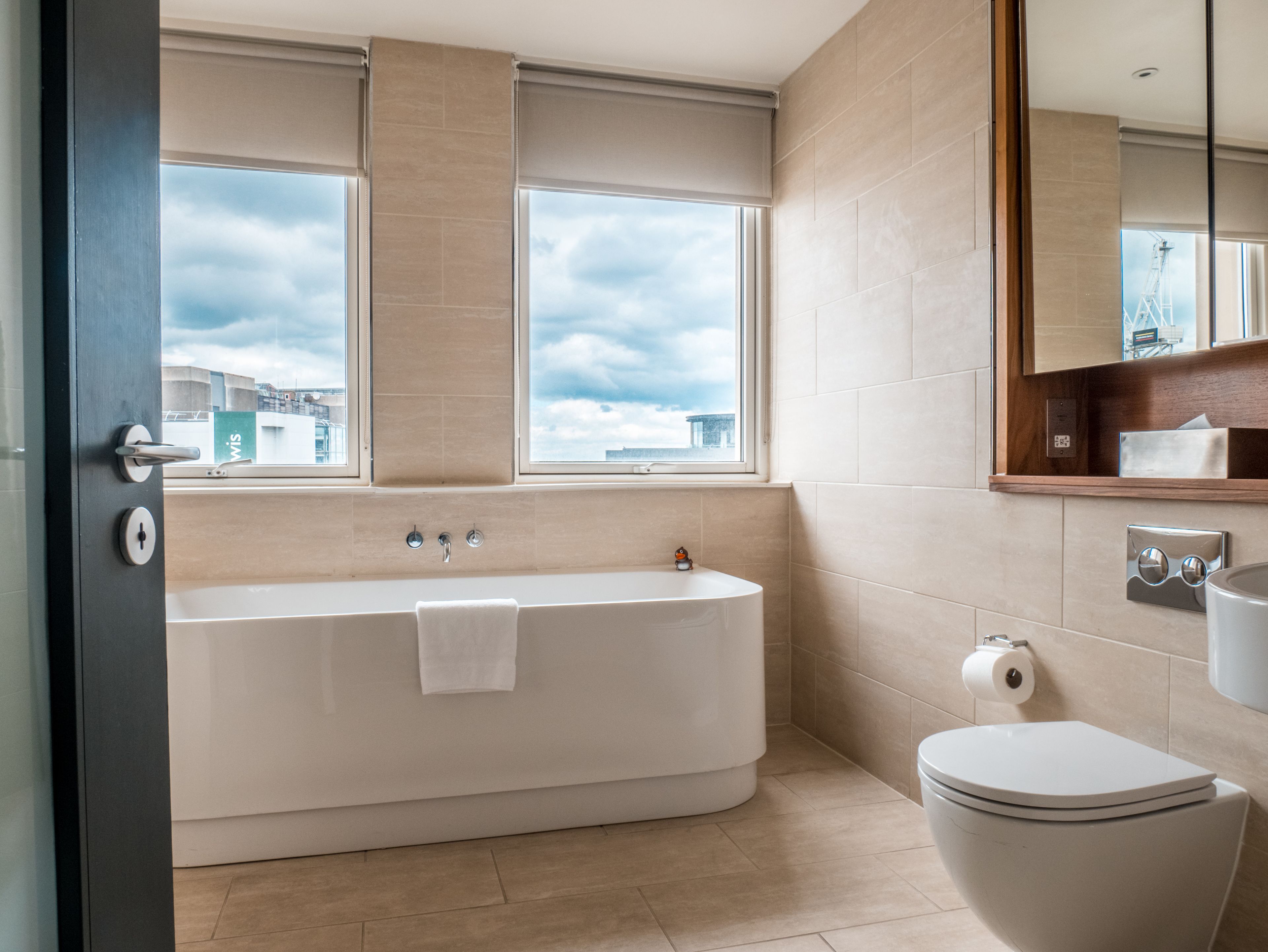 A modern bathroom with a bathtub, toilet, and a large window providing a view of the cityscape.