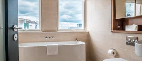 A modern bathroom with a bathtub, toilet, and a large window providing a view of the cityscape.