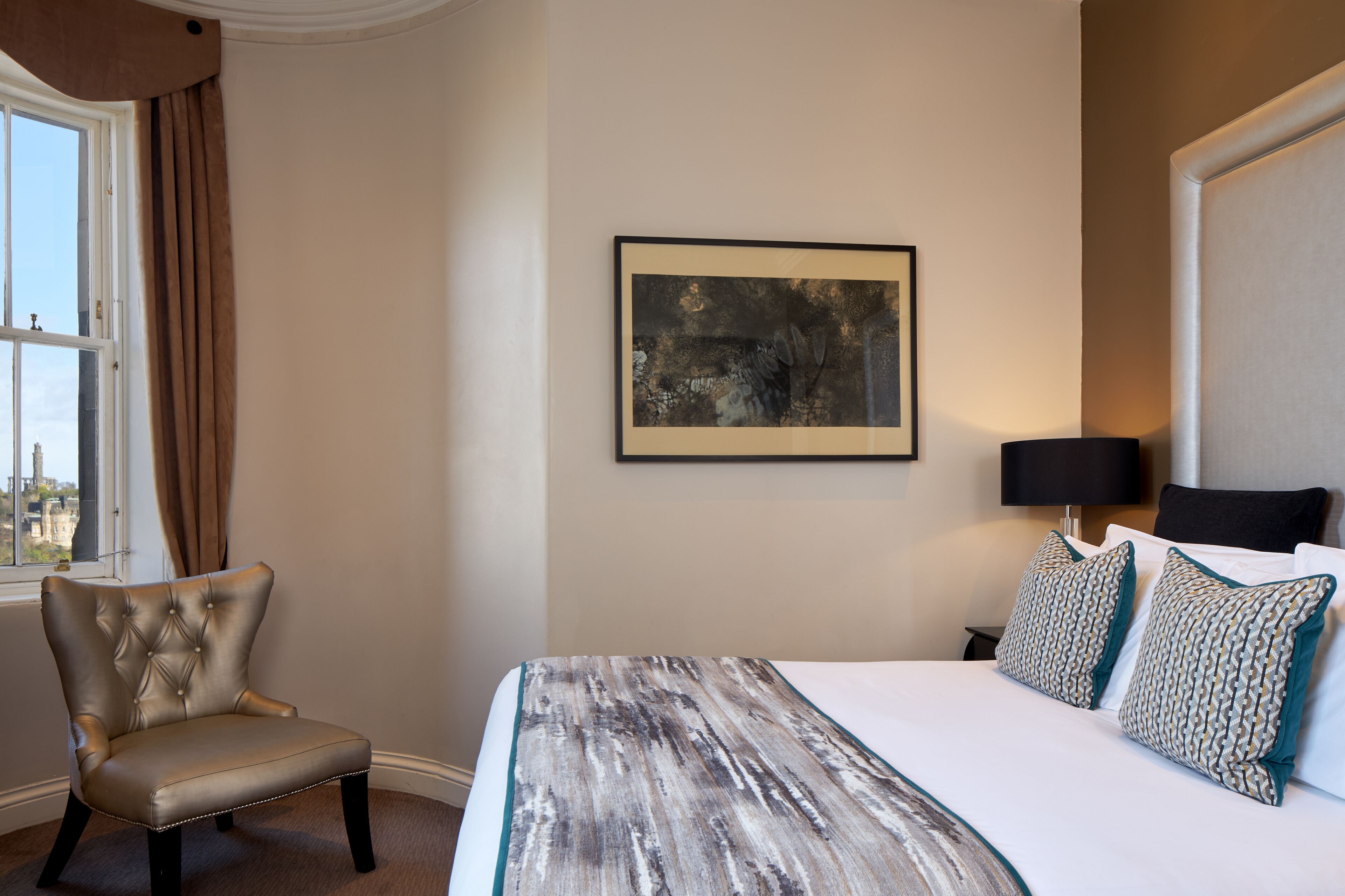 A hotel room with a bed, a chair, a framed artwork on the wall, and a window with curtains.