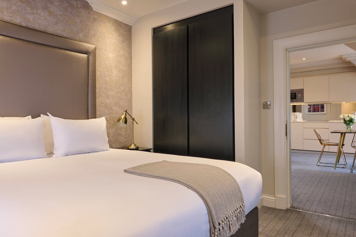 A hotel room with a large bed, a bedside lamp, a dark door, and a view into the kitchen area.