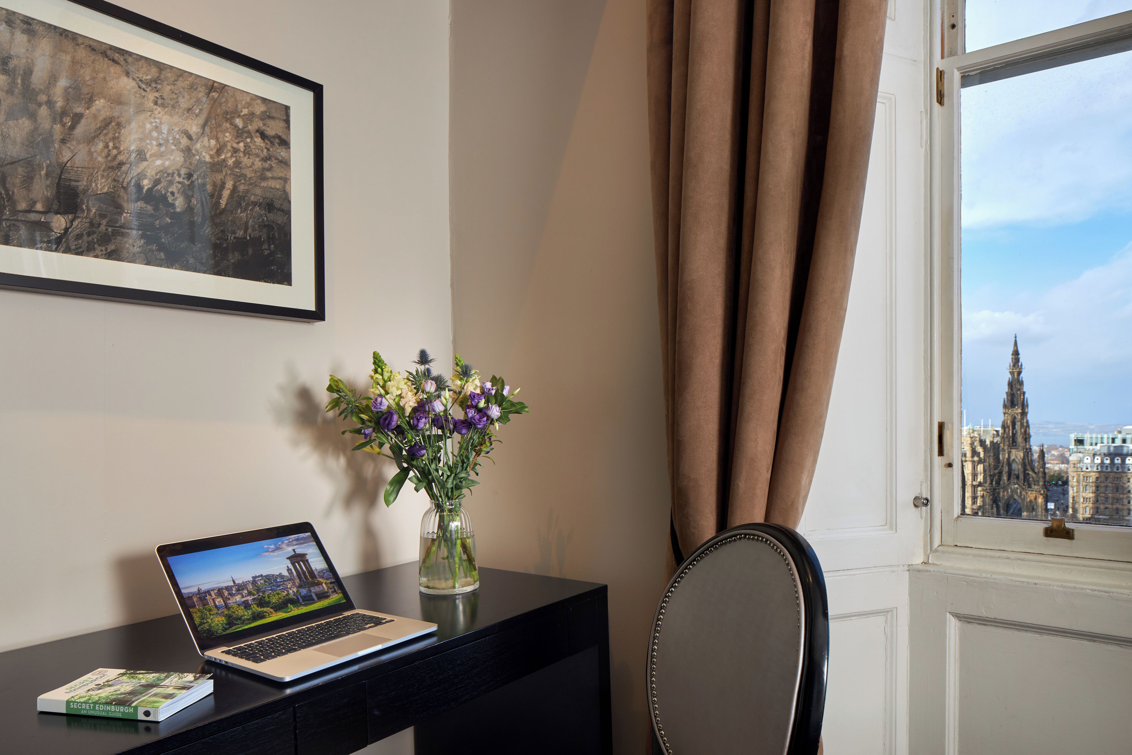 A laptop on a desk with a framed picture and a vase of flowers. A chair is next to the desk.