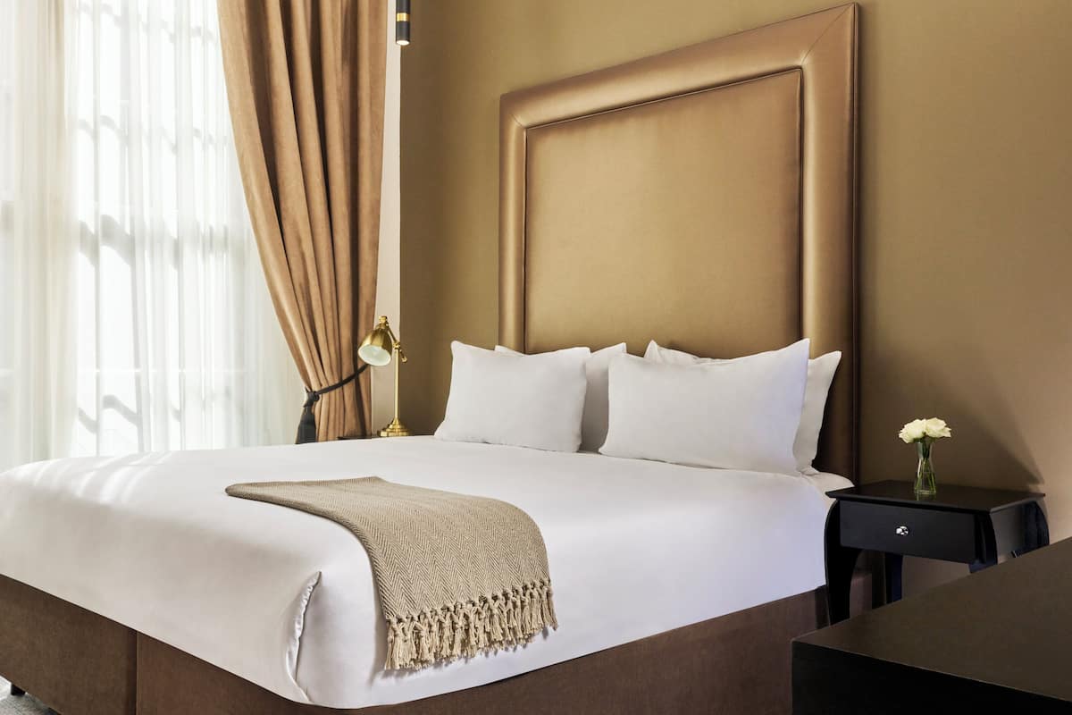 A neatly made bed with a beige throw blanket, a bedside lamp, and a vase with a flower on a nightstand.