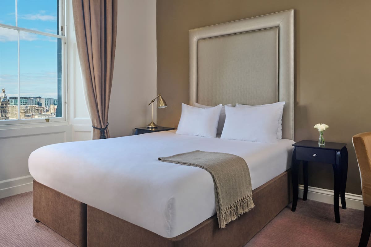A hotel room with a large bed, a nightstand, a desk, a chair, and a window with a city view.
