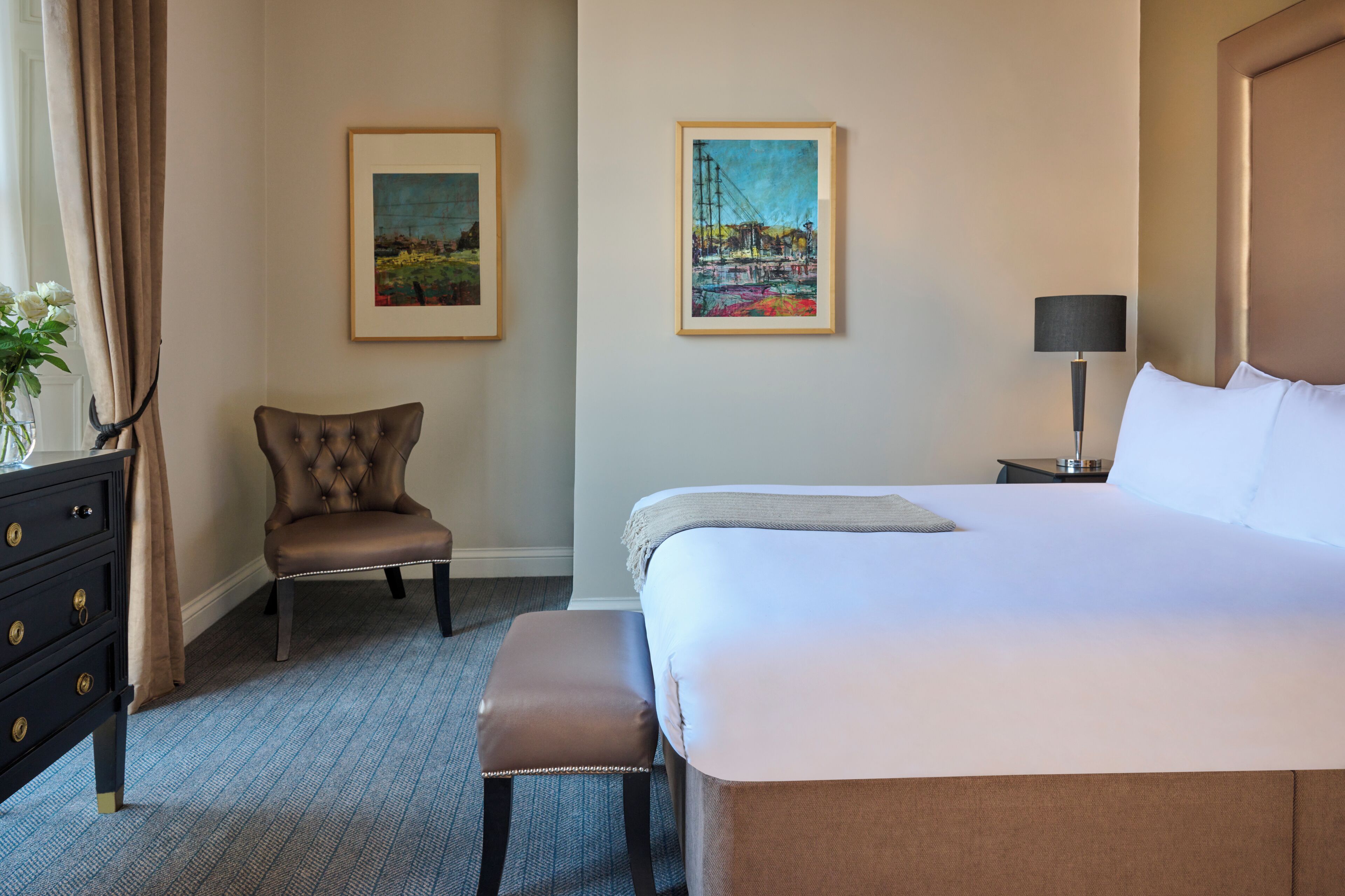 Habitación de hotel con una cama grande, una silla, un banco, un aparador y dos cuadros enmarcados en la pared.