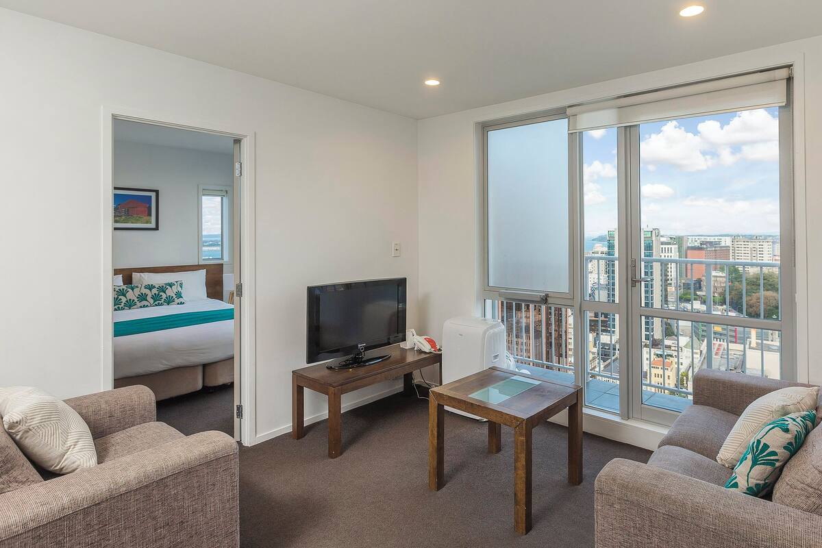 A modern living room with a flat-screen TV, a wooden coffee table, and two sofas. A bed with a blue and white bedspread is visible through an open door.