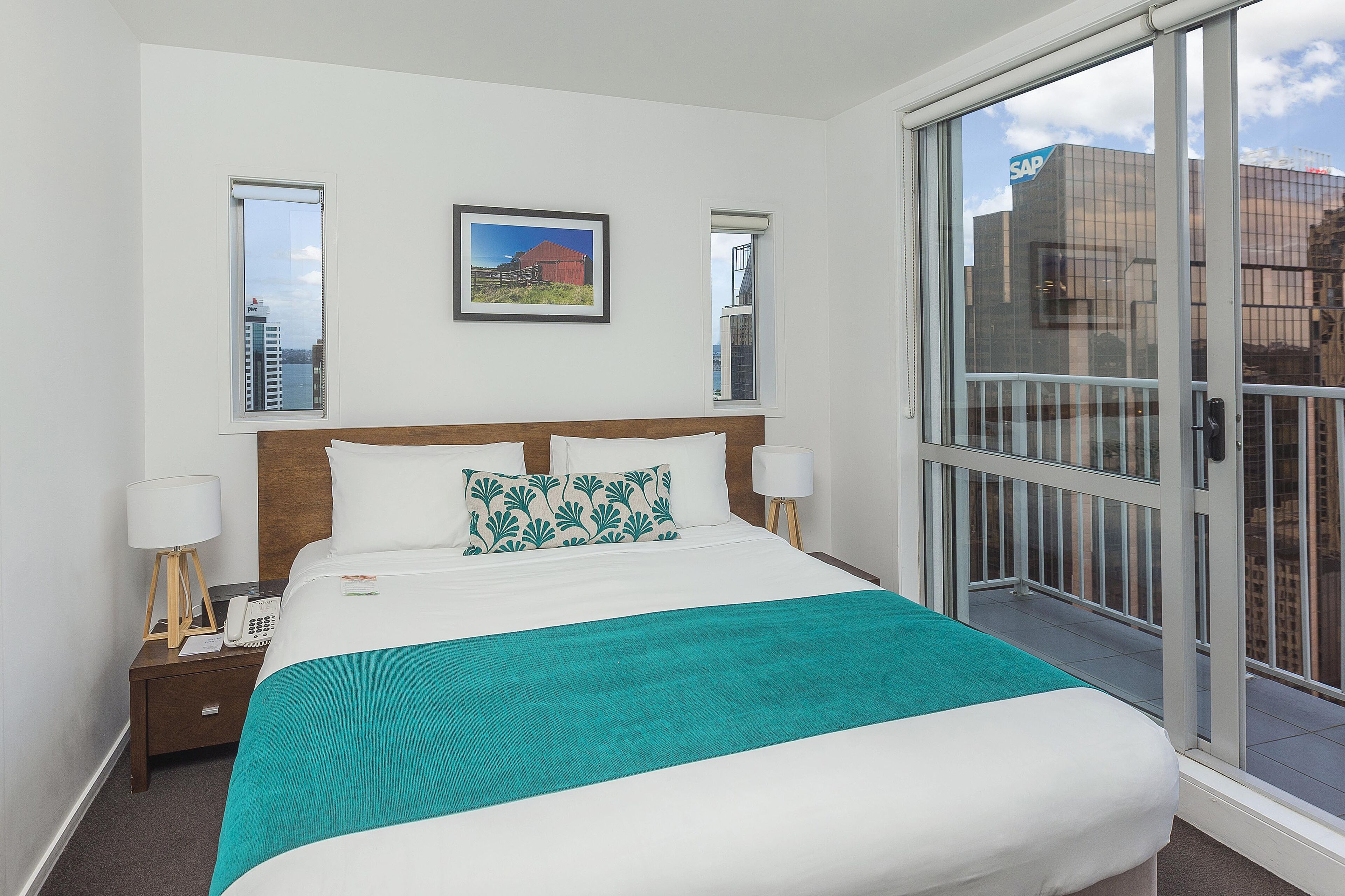 A hotel room with a bed, a nightstand with a lamp, a framed picture on the wall, and a view of the city through a large window.