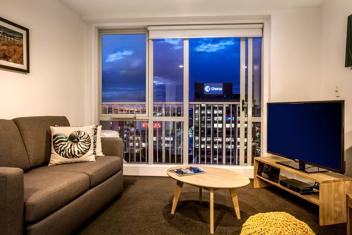 A modern living room with a sofa, a coffee table, a TV, and a large window with a city view.