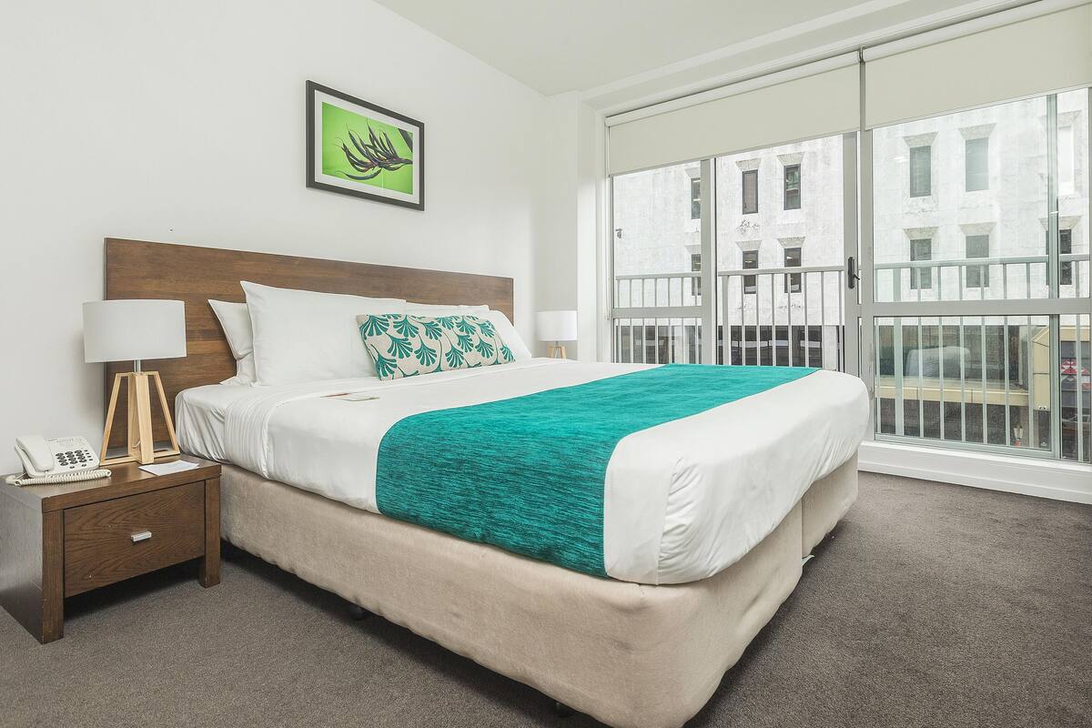 A hotel room with a bed, a nightstand, a telephone, and a window with a view of buildings.