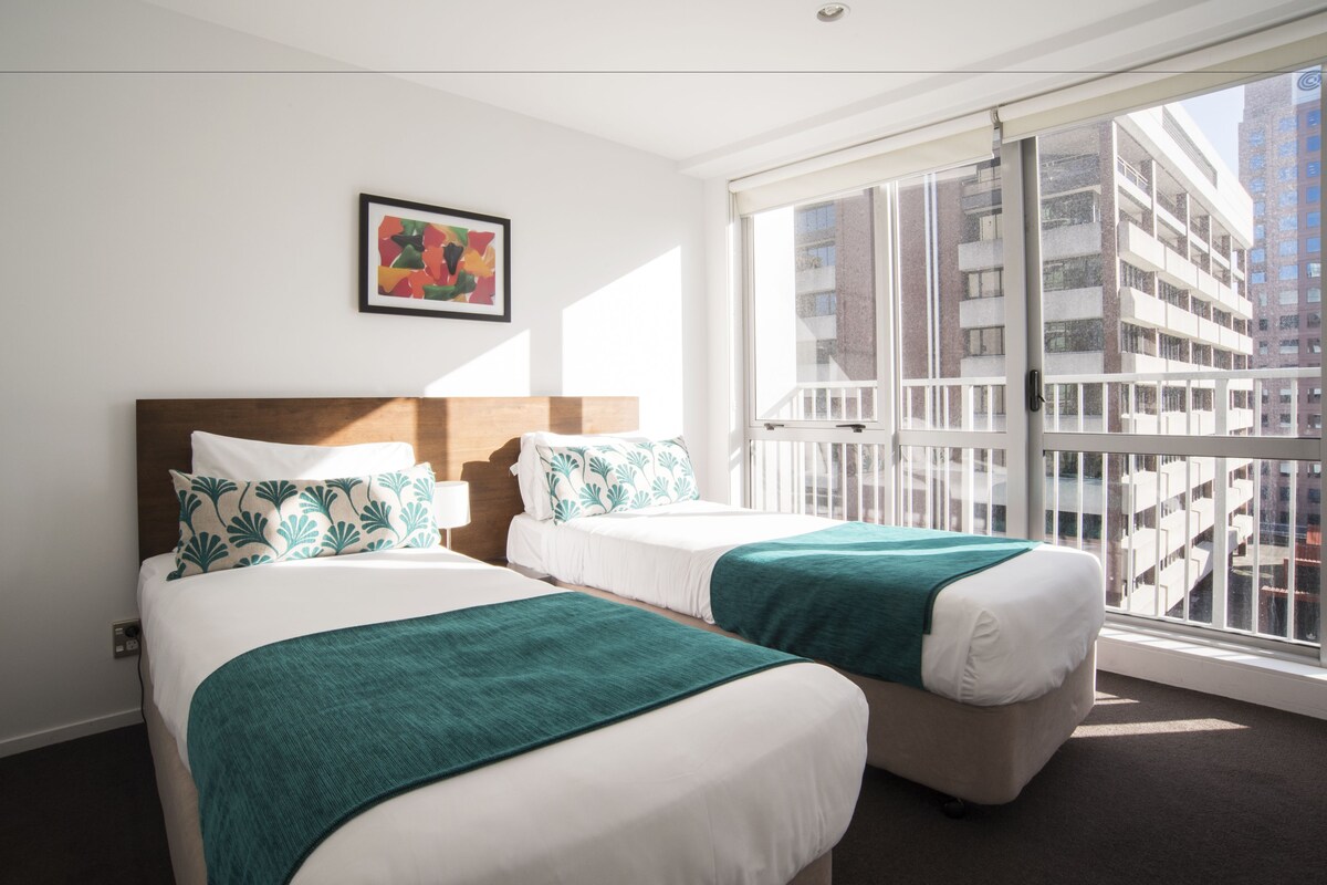 A hotel room with two beds, a large window with a view of buildings, and a framed picture on the wall.
