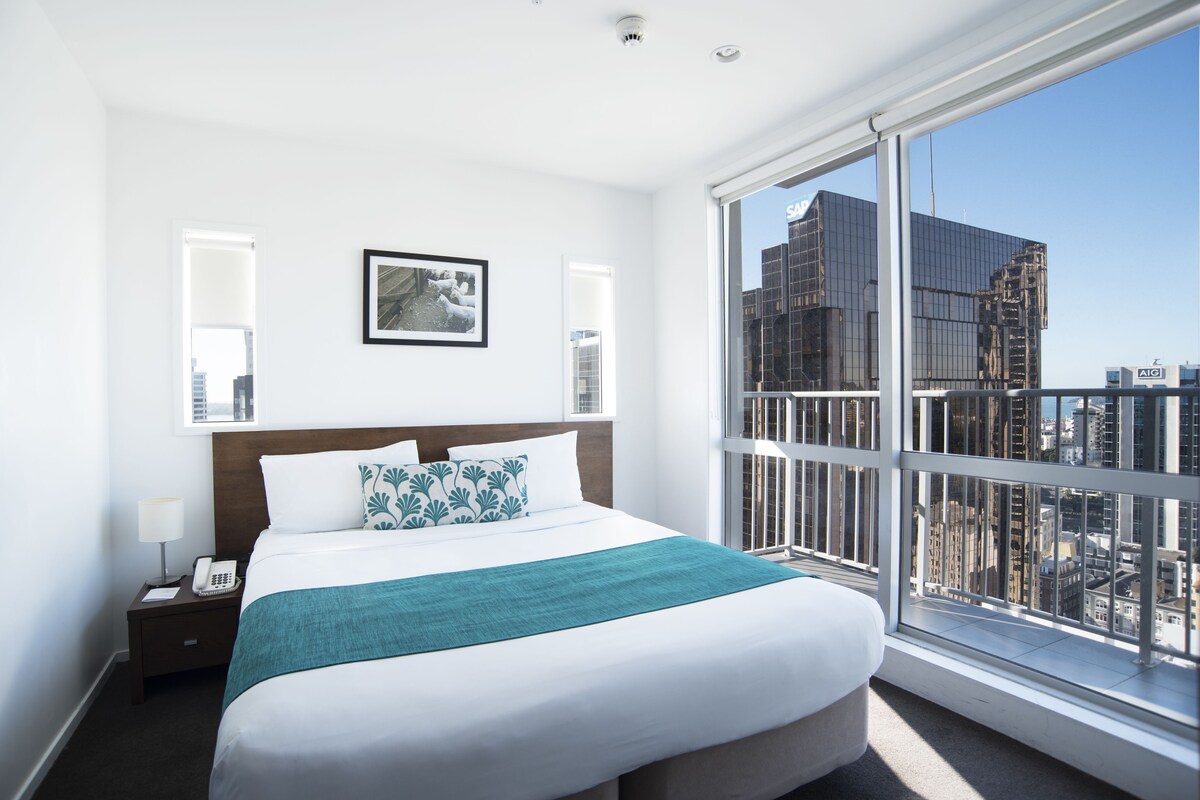 A hotel room with a large bed, a nightstand with a phone, a window with a city view, and a framed picture on the wall.
