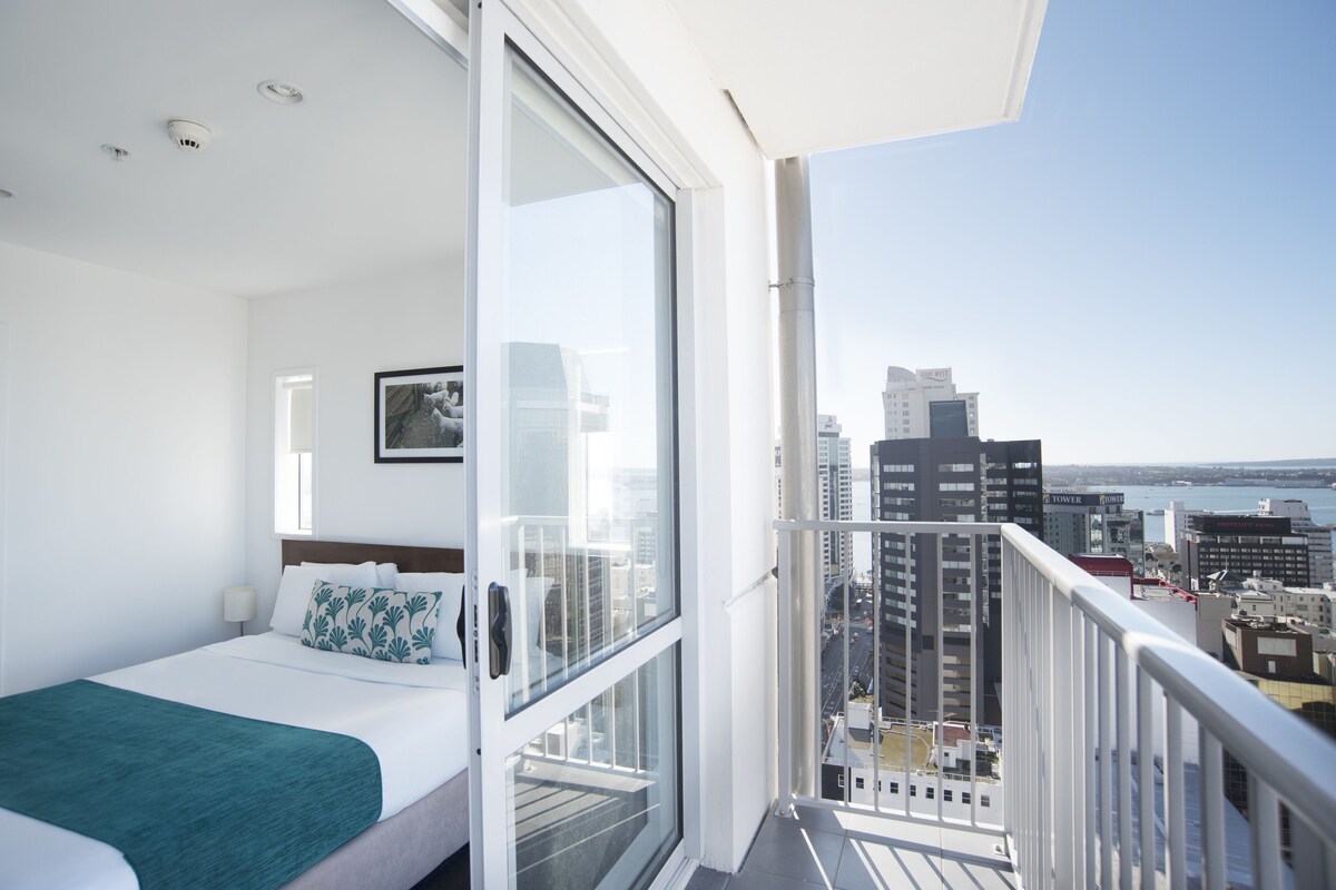 A hotel room with a bed, a window, and a balcony with a city view.