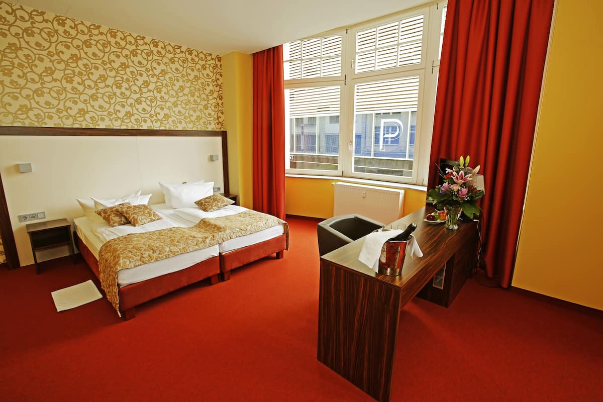 A hotel room with a double bed, a desk with a chair, a radiator, and a window with blinds.
