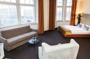 Habitación de hotel con cama, sofá, silla, una mesita y ventanas amplias con vistas a la ciudad.