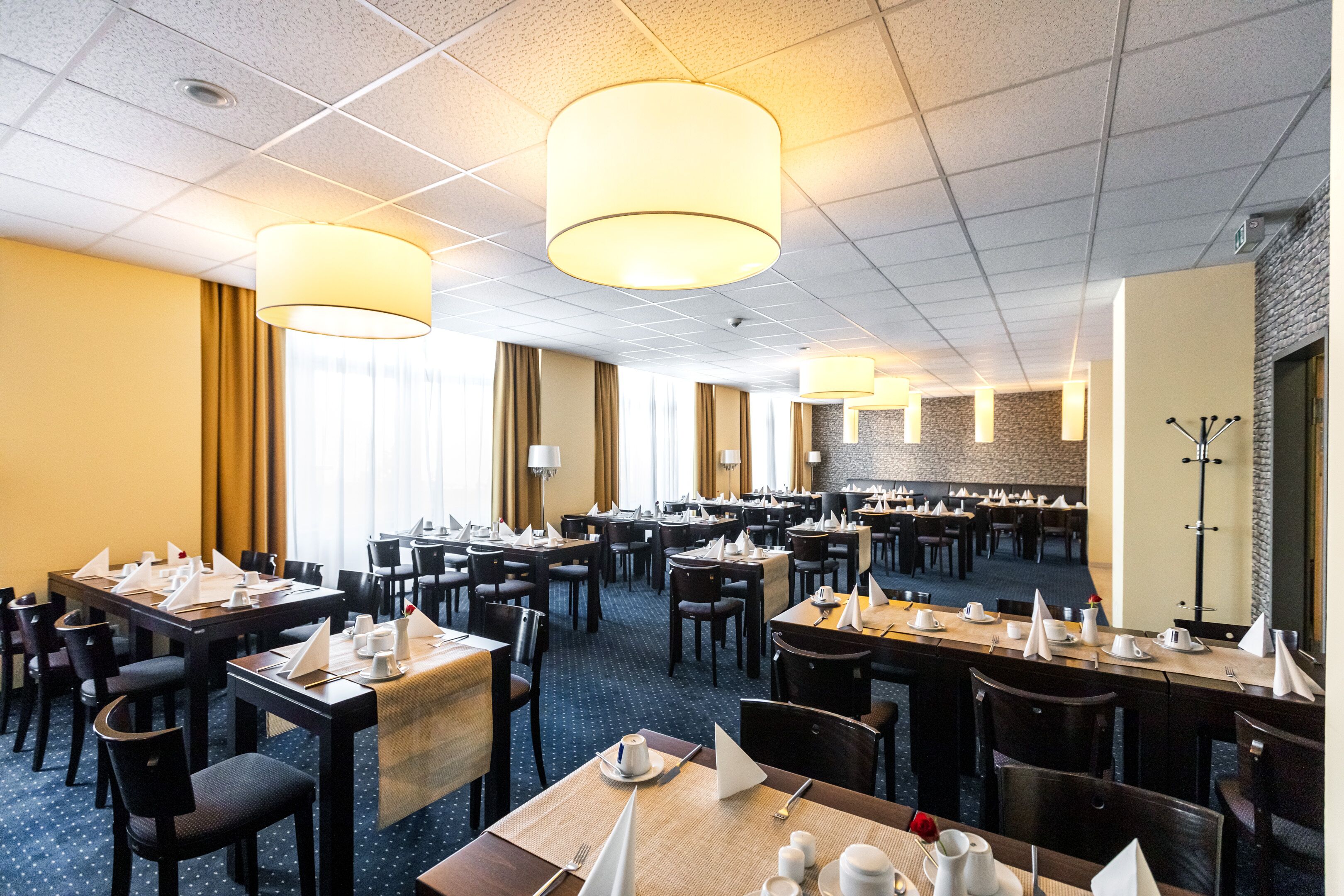 Un'area ristorazione in hotel con diversi tavoli e sedie, grandi lampadari a sospensione e una parete decorativa in pietra.