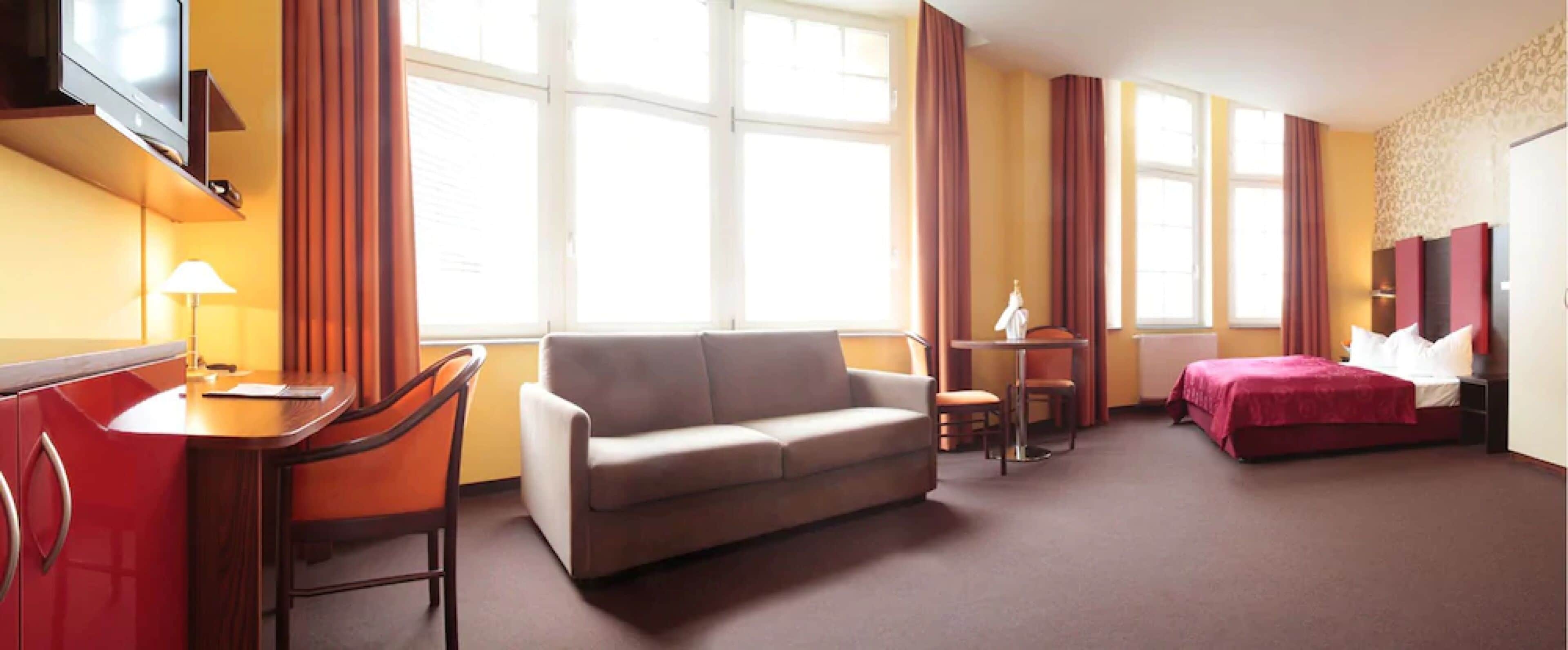 A hotel room with a desk, sofa, and bed. There are large windows with curtains, a TV mounted on the wall, and a lamp on the desk.
