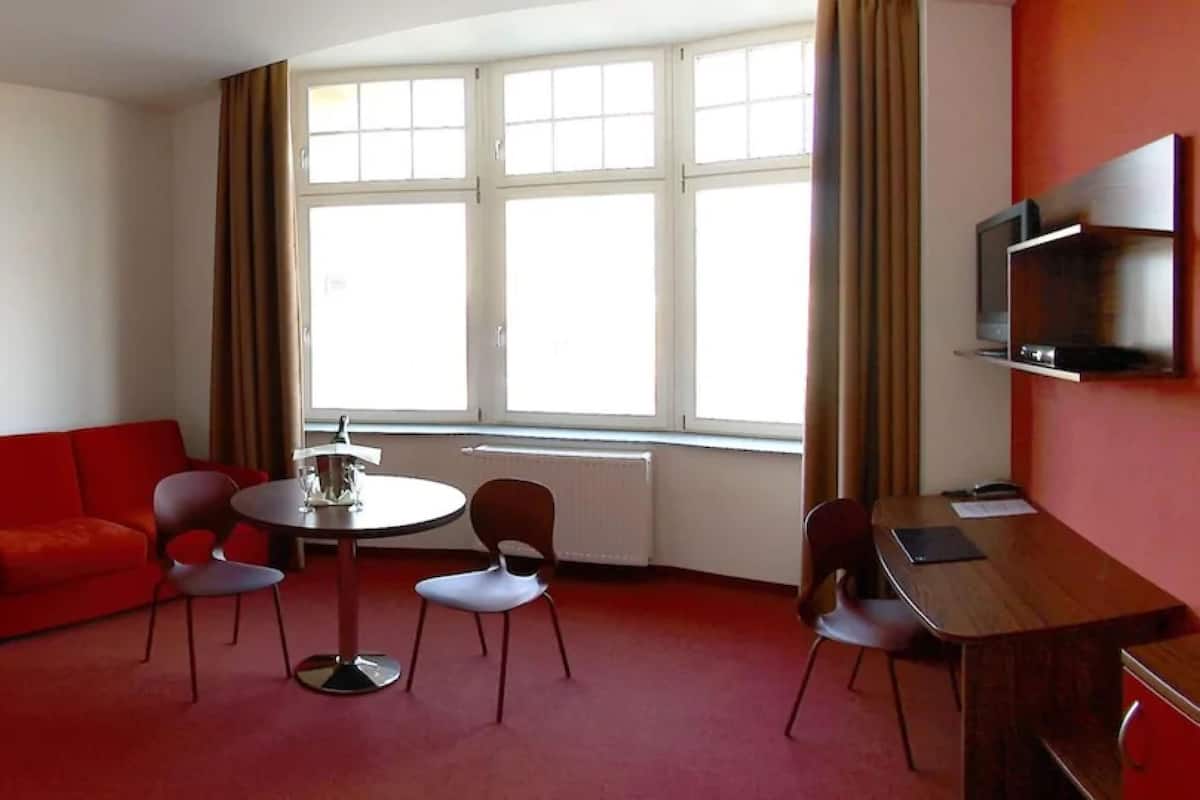 A hotel room with a red sofa, a round table with chairs, a TV mounted on the wall, and a window with curtains.